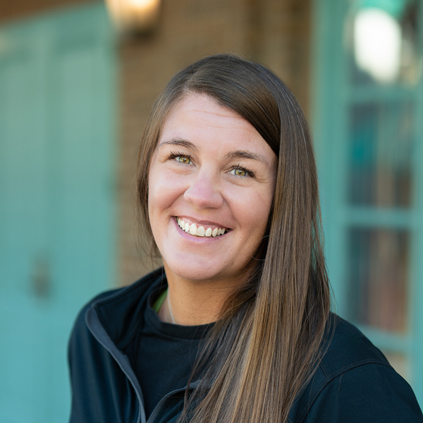 Nicole Hensley, Arrow Wellness Director at Arrow Senior Living Home Office, smiling in a professional outdoor headshot with long straight brown hair and softly blurred teal door, brick, and window details in the background.