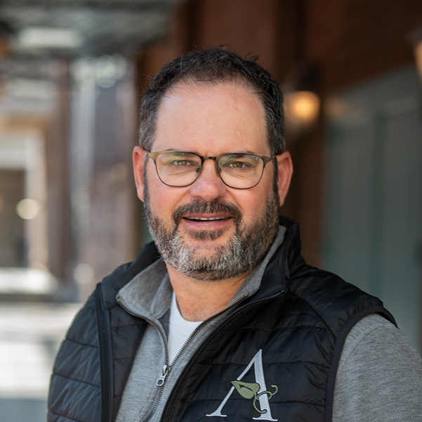 Tim Moore, Regional Director of Operations at Arrow Senior Living, smiling in a professional headshot, wearing glasses, a gray shirt, and a black vest, with a softly blurred outdoor background featuring brick and architectural details.