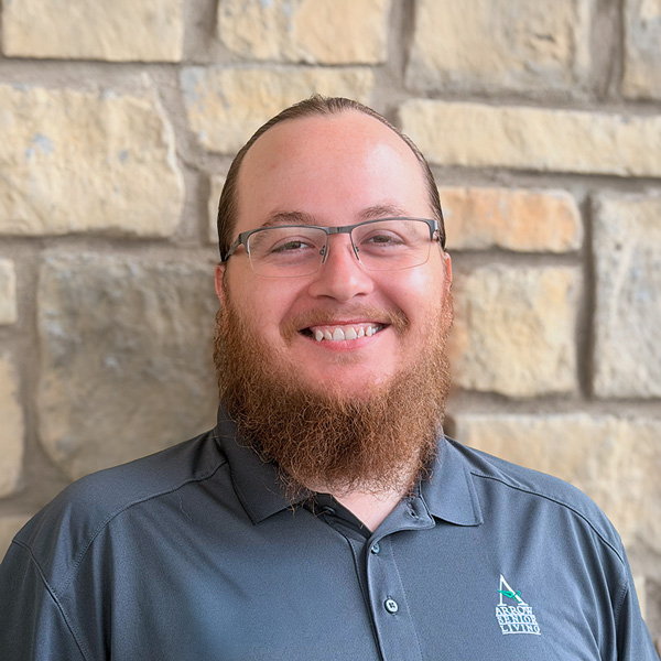 Tom Reynolds, Plant Operations Director - Travel Team at Arrow Senior Living Home Office, smiling in a professional headshot, wearing glasses and a gray polo, with a stone wall background.