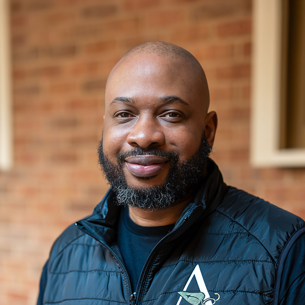 Aaron Blevins, Regional Wellness Coordinator at Arrow Senior Living Home Office, looking at the camera in a professional outdoor headshot with a bald head, full beard, black vest, and a softly blurred brick background.