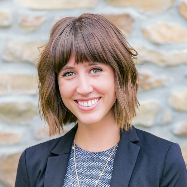 Amber Moore, Managing Director of Human Resources at Arrow Senior Living Home Office, smiling in a professional headshot with a short brown bob, bangs, a black blazer, layered necklaces, and a light stone wall background.