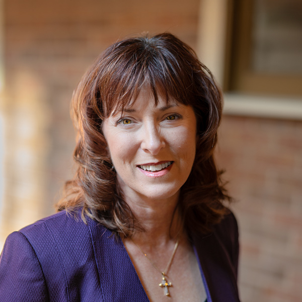 Becky Kaufmann, Managing Director of Wellness at Arrow Senior Living Home Office, smiling in a professional outdoor headshot with auburn shoulder-length hair, a purple blazer, a cross necklace, and a softly blurred brick background.