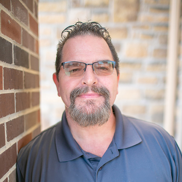 Bill Crawley, Regional Plant Operations Coordinator at Arrow Senior Living Home Office, looking at the camera in a professional headshot with glasses, short curly dark hair, a salt-and-pepper goatee, a gray polo shirt, and a softly blurred brick and stone background.