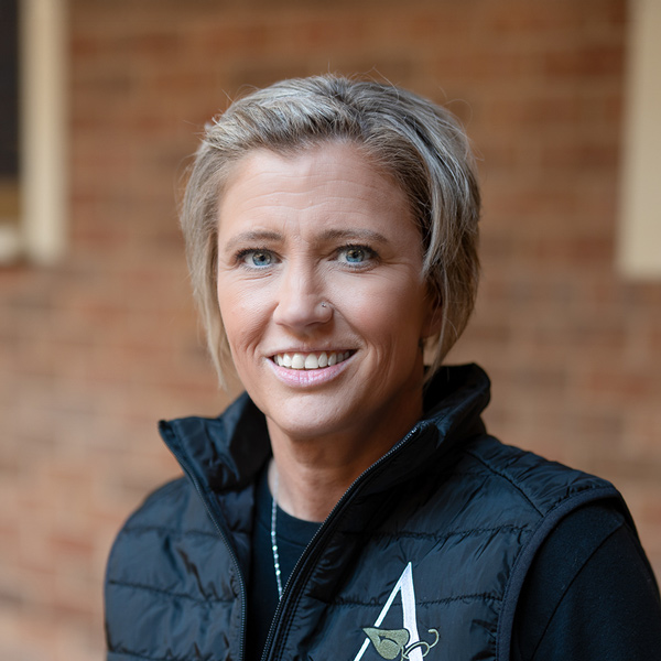 Christy Kay, Regional Director of Wellness at Arrow Senior Living Home Office, smiling in a professional outdoor headshot with short blonde hair, a black vest, and a softly blurred brick background.