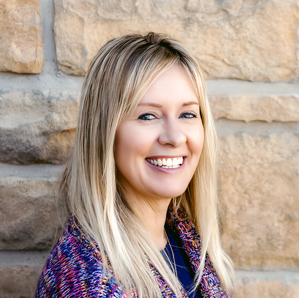 Tracy Haarmann, Regional Executive Director at Arrow Senior Living, smiling in a colorful sweater with long blonde hair, standing beside a tan stone wall.