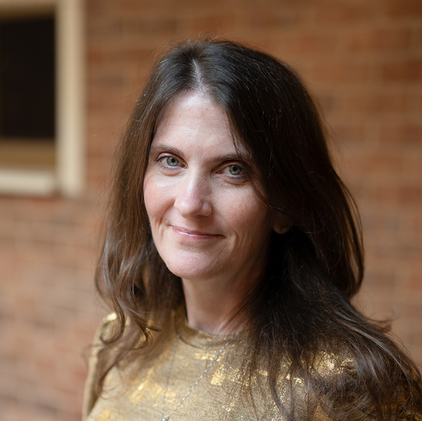 Holly Belanger, Senior Regional Wellness Coordinator at Arrow Senior Living Home Office, looking at the camera in a professional outdoor headshot with long dark brown hair, a gold top, and a softly blurred brick background.