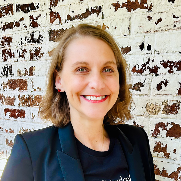 Jen Johnmeyer, Director of Brand Experience at Arrow Senior Living Home Office, smiling in a professional headshot with short wavy light brown hair, a black blazer, bright lipstick, and a white painted brick wall background.