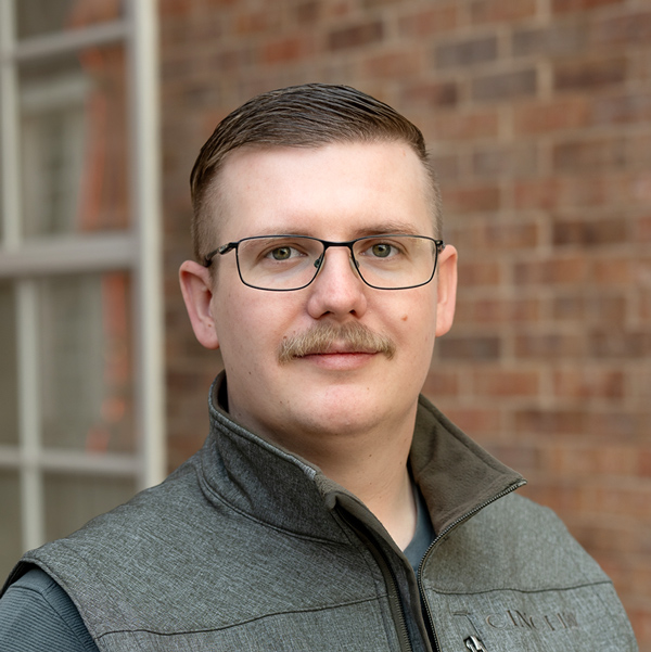 Justin Roberts, Senior Regional Director of Plant Operations at Arrow Senior Living Home Office, looking at the camera in a professional outdoor headshot with short neatly combed hair, glasses, a mustache, a gray vest, and a softly blurred brick background.