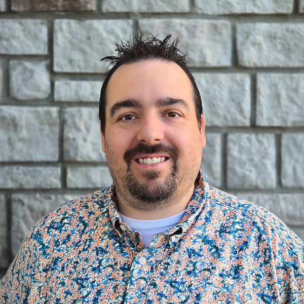 Justin Stewart, Arrow Senior Living Director at Arrow Senior Living Home Office, smiling in a professional headshot with short dark spiked hair, a goatee, a colorful floral button-up shirt, and a gray stone wall background.