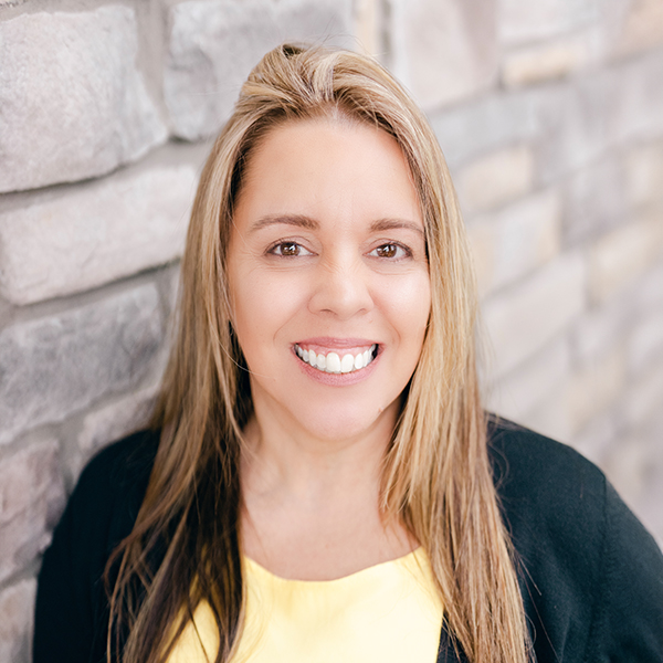 Katie Crespo, Arrow Senior Living Director at Arrow Senior Living Home Office, smiling in a professional headshot with long highlighted hair, a yellow top, and a softly blurred light stone wall background.