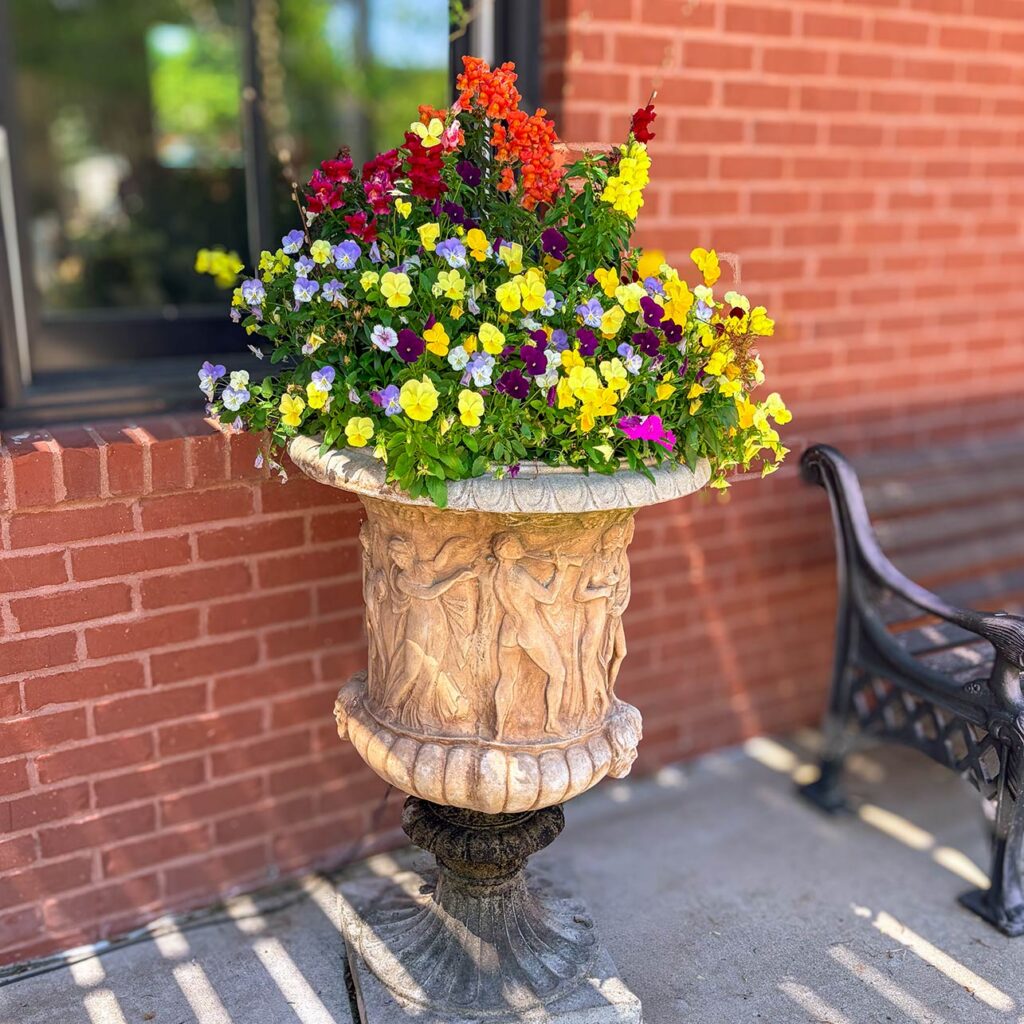 An elegant stone planter full of colorful spring flowers rests beside a bench at Arrow Senior Living home office.