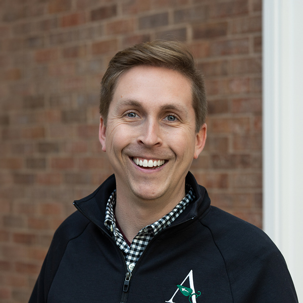 Rob Cain, Vice President of Compliance at Arrow Senior Living Home Office, smiling in a professional outdoor headshot with short light brown hair, a black quarter-zip jacket over a checkered shirt, and a softly blurred brick background.