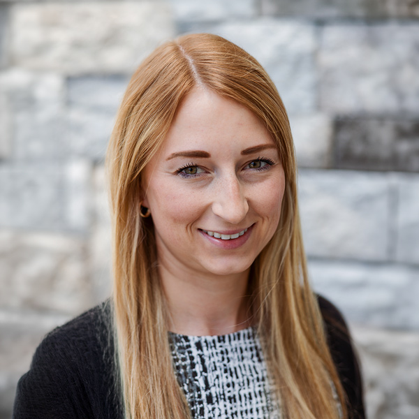 Stefanie Finley, Portfolio Director at Arrow Senior Living Home Office, smiling in a professional headshot with long straight strawberry blonde hair, a patterned black and white top, and a softly blurred gray stone wall background.