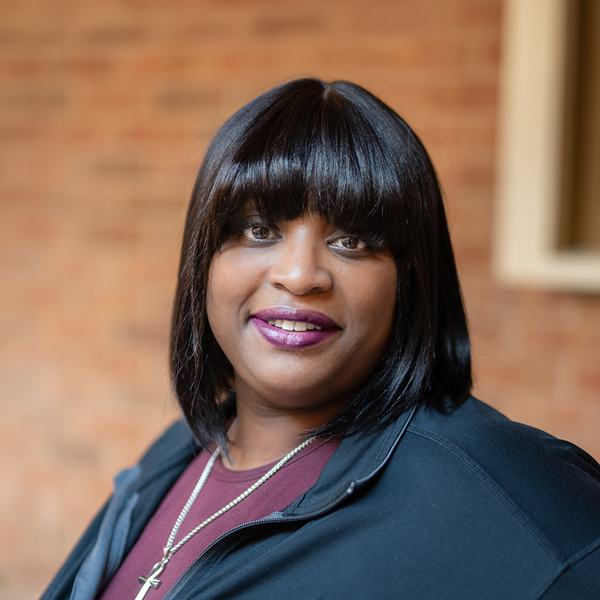 Octavia “Tay” Lawler-Thomas, Arrow Wellness Director at Arrow Senior Living Home Office, looking at the camera in a professional outdoor headshot with a sleek black bob and bangs, a black jacket, a berry-colored top, and a softly blurred brick background.