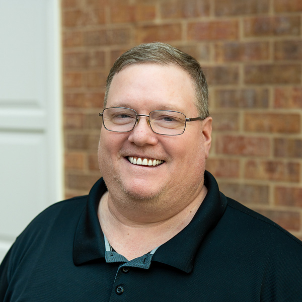 Tim Harmon, Regional Plant Operations Coordinator at Arrow Senior Living Home Office, smiling in a professional outdoor headshot with short light brown hair, glasses, a black polo shirt, and a softly blurred brick background.