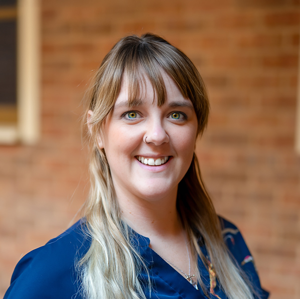 Tori O’Hagon, Regional Wellness Coordinator at Arrow Senior Living Home Office, smiling in a professional outdoor headshot with blonde hair and bangs, a blue top, a small nose ring, and a softly blurred brick background.