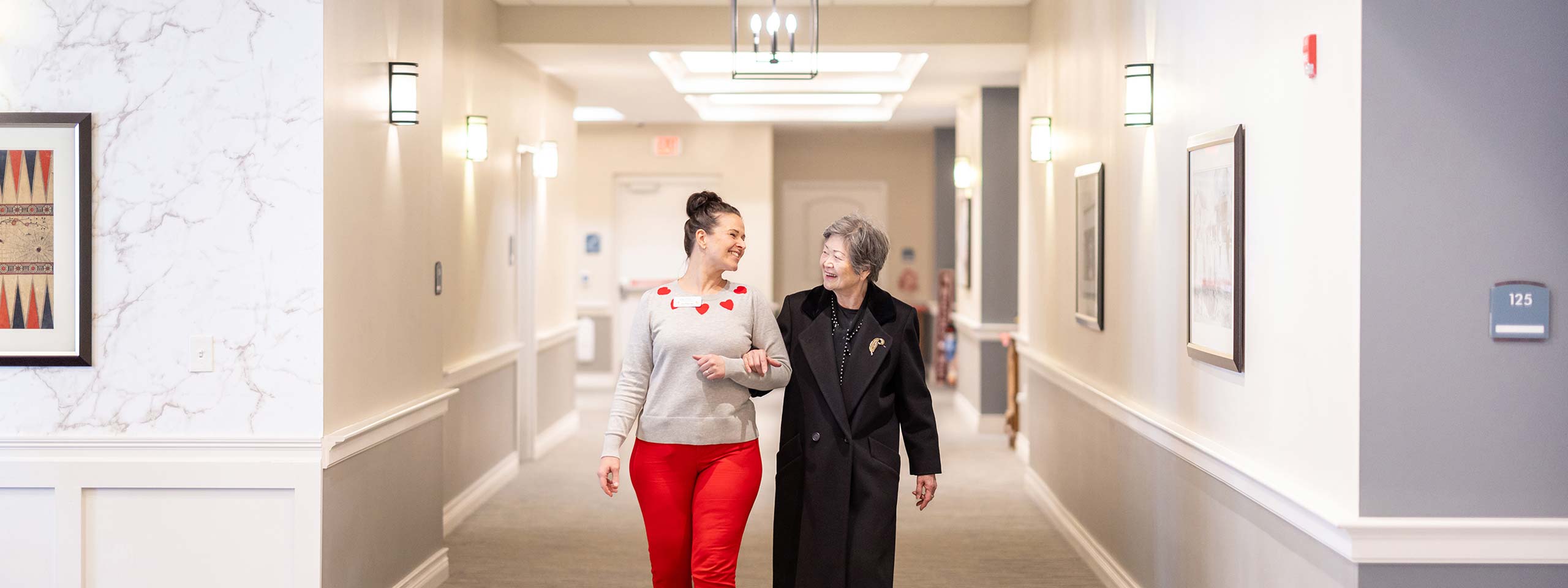 Happy young woman in a gray sweater with red hearts walks arm-in-arm with a senior resident in a black coat, both smiling as they stroll through a brightly lit hallway.