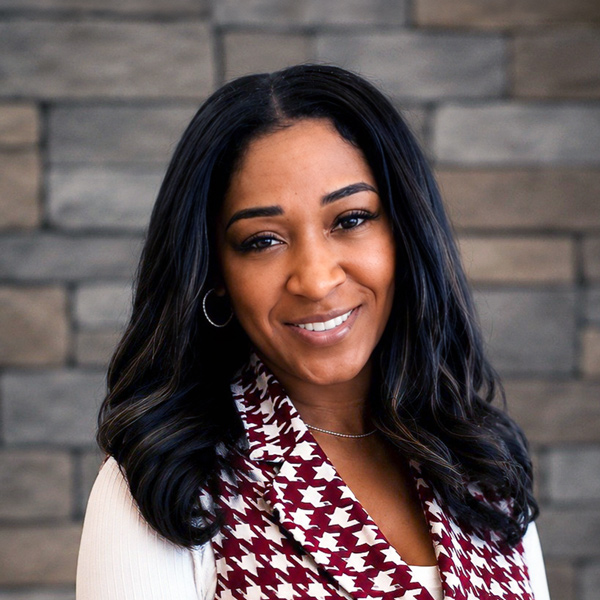 Crystal Parrish, Senior Managing Director of Wellness at Arrow Senior Living Home Office, smiling in a professional headshot with long dark wavy hair, hoop earrings, a patterned scarf, and a stacked gray stone wall background.