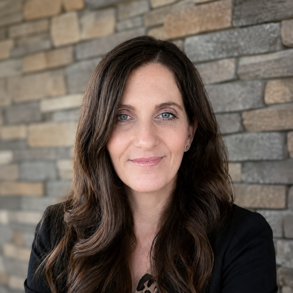 Holly Belanger, Senior Regional Wellness Coordinator at Arrow Senior Living Home Office, looking at the camera in a professional headshot with long dark wavy hair, a black blazer, and a stacked stone wall background.