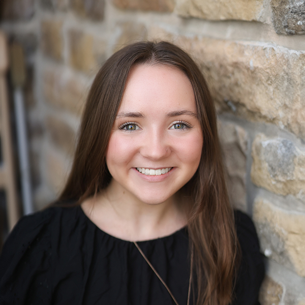 Paige Amsberry, Regional Operations Specialist at Arrow Senior Living Home Office, smiling in a professional headshot with long straight brown hair, a black blouse, a pendant necklace, and a softly blurred stone wall background.