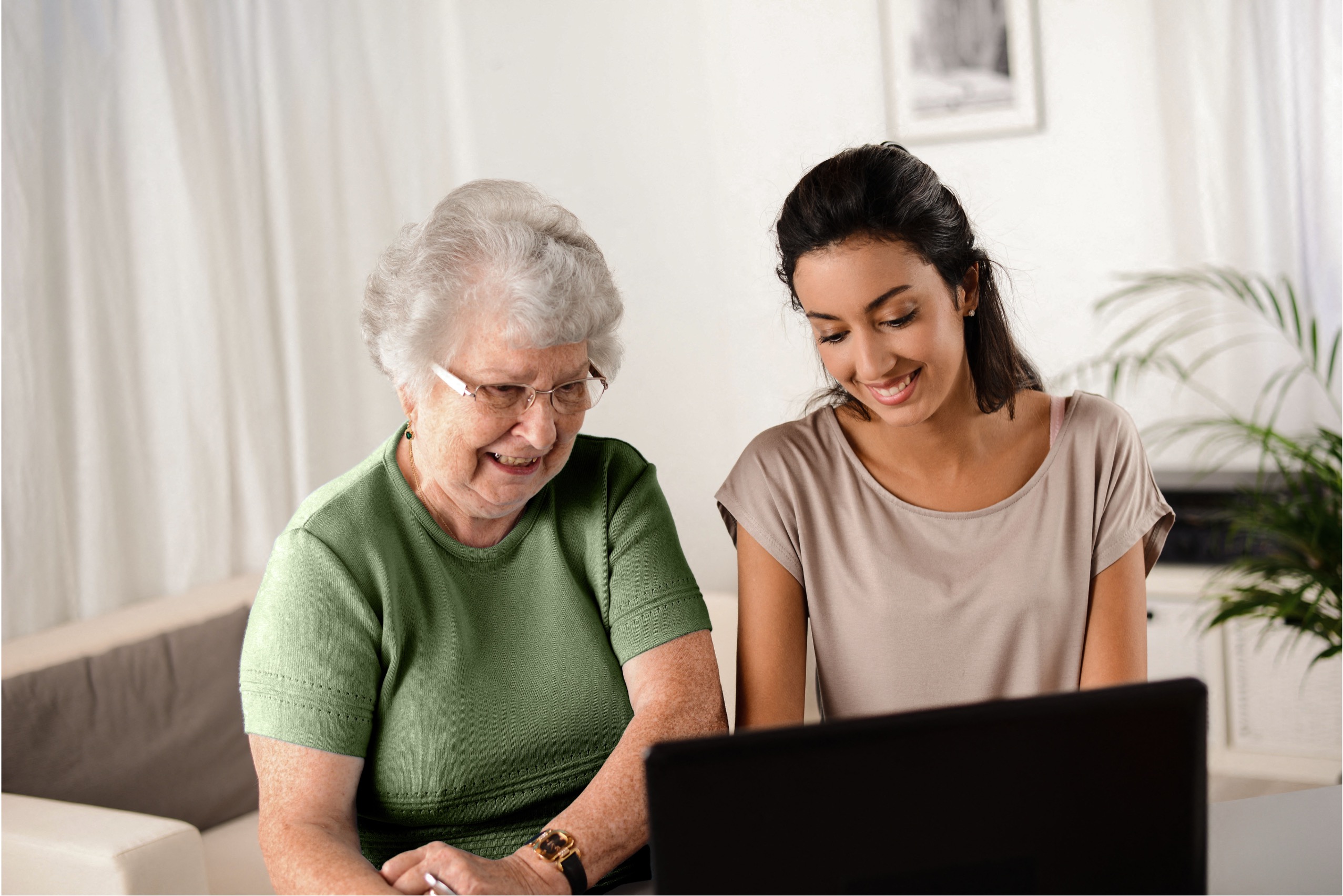 Senior living resident and team member smiling while looking at a laptop together in a bright community space.