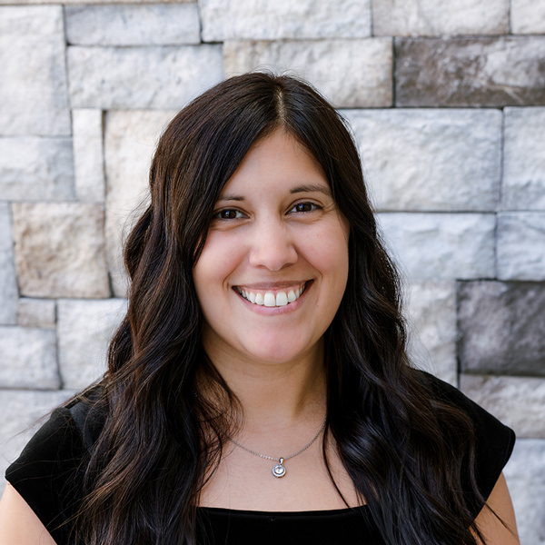Lauren Snelling, Arrow Dining Room Manager for the Kansas City area at Arrow Senior Living Home Office, smiling in a professional headshot with long dark wavy hair, a black top, a pendant necklace, and a gray stone wall background.