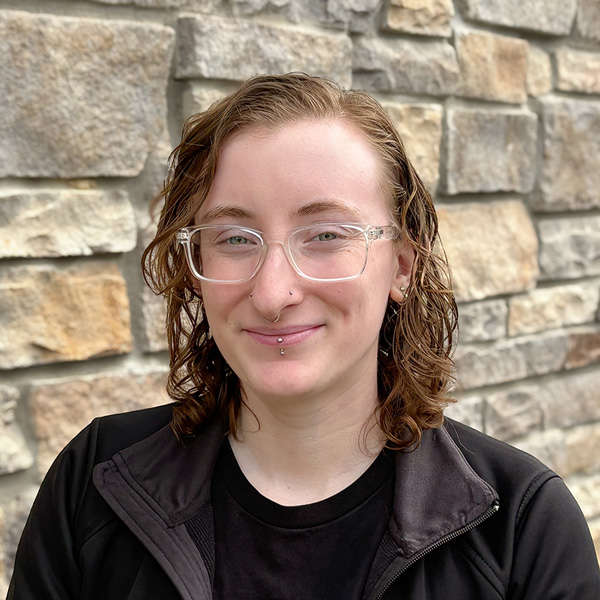 Olivia “Liv” Rainey, Arrow Dining Room Manager at Arrow Senior Living Home Office, smiling in a professional headshot with short wavy light brown hair, clear glasses, facial piercings, and a stacked stone wall background.