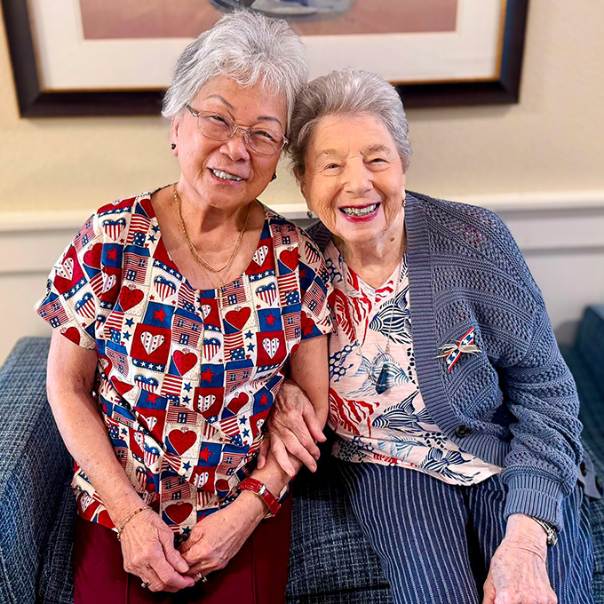 Two senior women wearing patriotic clothing and accessories smile brightly together during a community event.