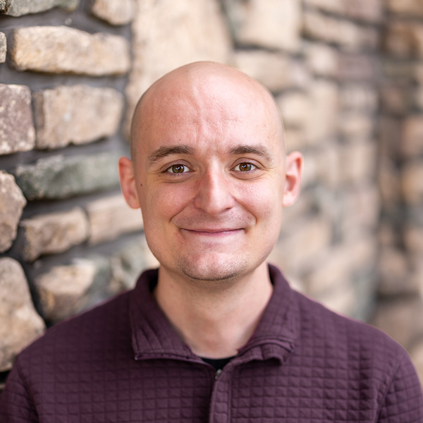 Austin Marsh, Resident Experience Coordinator at Arrow Senior Living Home Office, smiling in a professional headshot with a bald head, a burgundy quarter-zip shirt, and a softly blurred stone wall background.