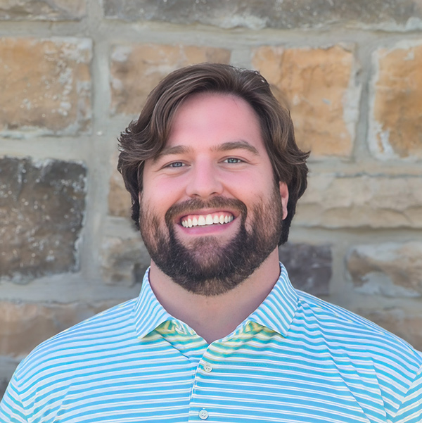 Ben Pinter, Arrow Senior Living Director at Arrow Senior Living Home Office, smiling in a professional headshot with wavy light brown hair, a full beard, a blue striped polo shirt, and a tan stone wall background.