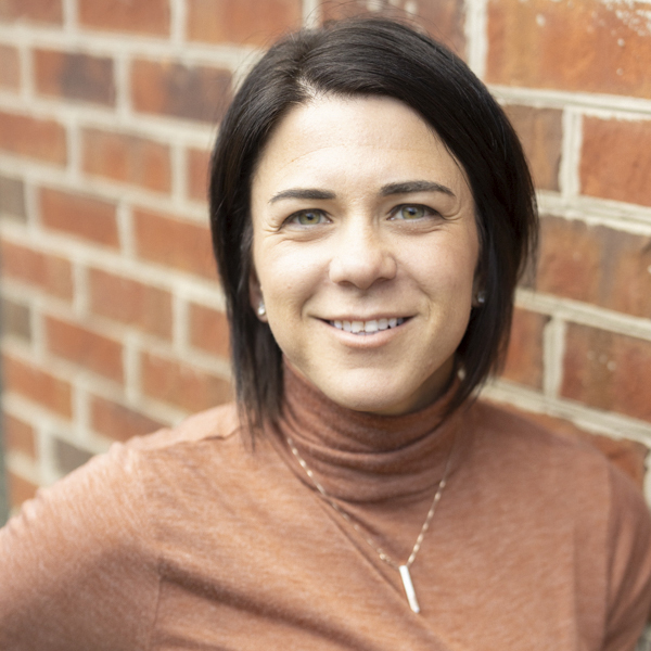 Karra Belza, Regional Executive Director for Arrow Senior Living, smiling in a professional headshot with short dark hair, a rust turtleneck top, a pendant necklace, and a softly blurred red brick wall background.