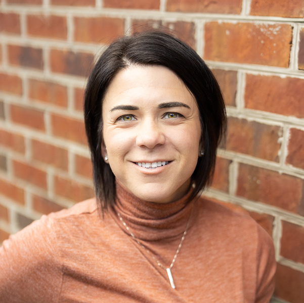 Karra Belza, Regional Executive Director for Arrow Senior Living, smiling in a professional headshot with short dark hair, a rust turtleneck top, a pendant necklace, and a softly blurred red brick wall background.