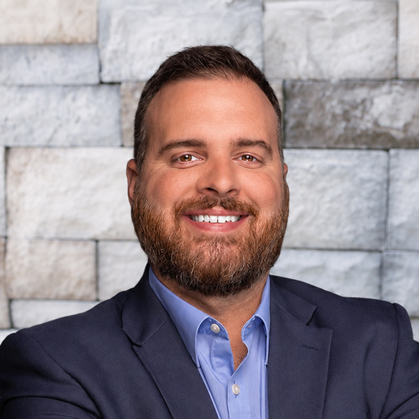 Ryan Potts, Corporate Counsel at Arrow Senior Living Home Office, smiling in a professional headshot with short dark hair, a full beard, a navy blazer, and a light gray stone wall background.