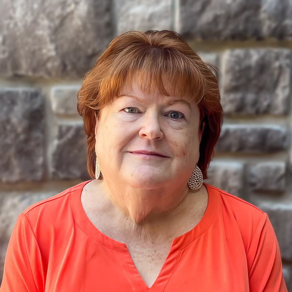 Renee Shea, Engagement Specialist at Arrow Senior Living Home Office, looking at the camera in a professional headshot with short auburn hair, drop earrings, a coral blouse, and a gray stone wall background.