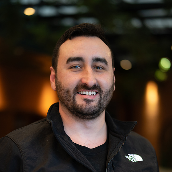 Dylan Hollister-Graham, Regional Executive Director at Arrow Senior Living Home Office, smiling in a professional headshot with short dark hair, a trimmed beard, a black vest, and a softly blurred indoor background.
