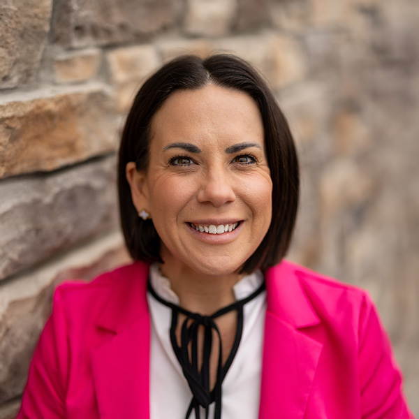 Heather Brenneman, Arrow Senior Living Director at Arrow Senior Living Home Office, smiling in a professional headshot with short dark hair, a bright pink blazer, and a softly blurred stone wall background.